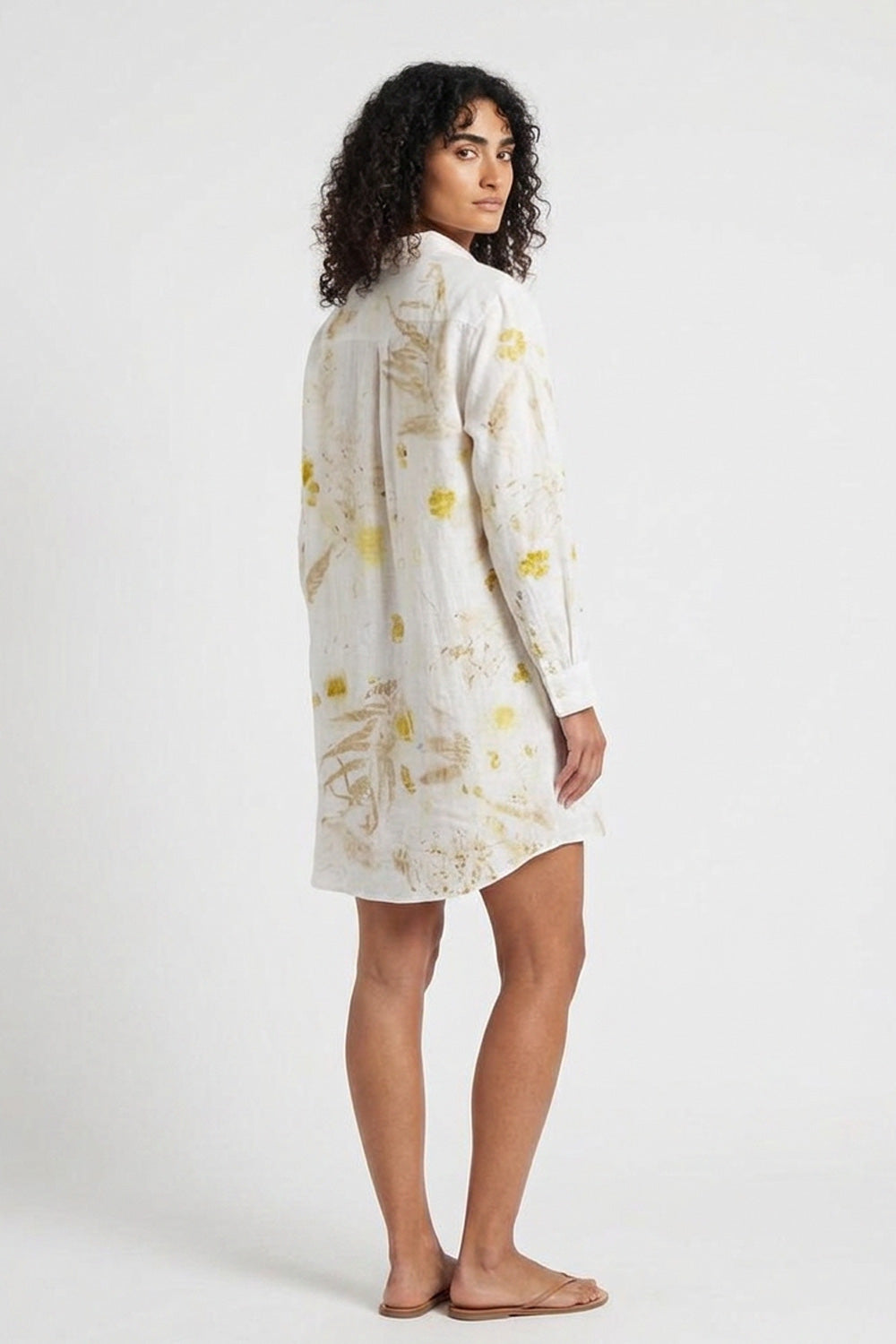 A woman modeling a long-sleeve white shirt dress with a yellow and gold abstract floral pattern, shown from a side-back angle against a plain white studio background.