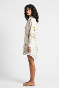 A woman stands in profile wearing a long-sleeve white shirt dress with a yellow and brown floral print, photographed against a plain light gray studio background.