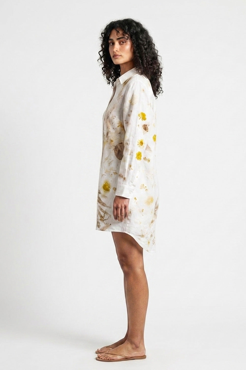 A woman stands in profile wearing a long-sleeve white shirt dress with a yellow and brown floral print, photographed against a plain light gray studio background.