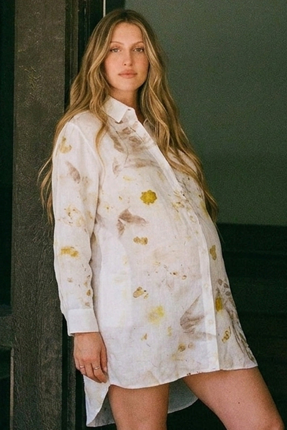 A pregnant woman wearing a white long-sleeve button-up shirt dress with a yellow and brown floral eco-print, leaning against a dark wooden pillar.