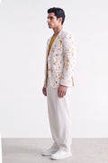 A man stands in profile wearing a white blazer with a yellow floral pattern, a mustard yellow shirt, white trousers, and white sneakers against a plain white studio background.