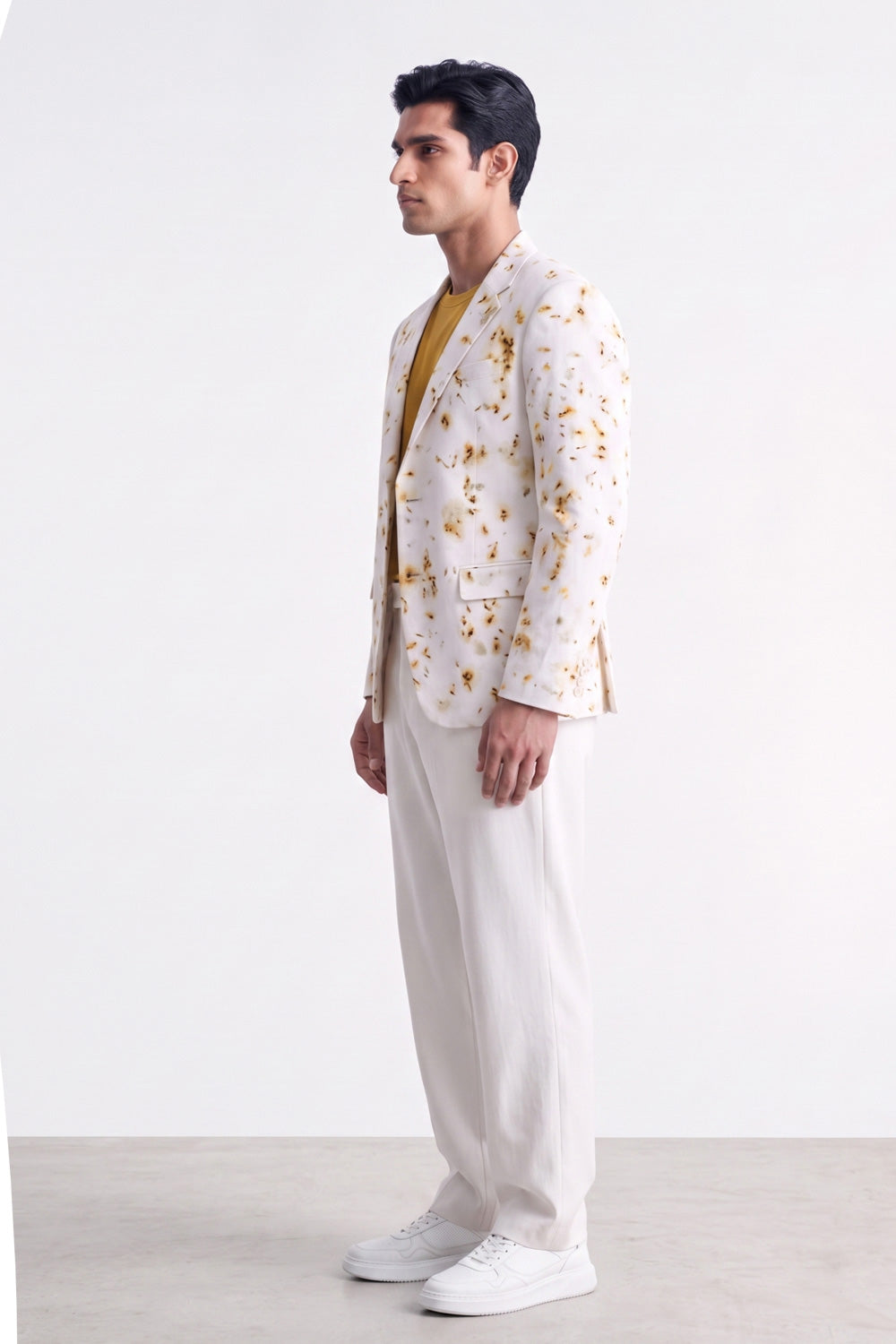 A man stands in profile wearing a white blazer with a yellow floral pattern, a mustard yellow shirt, white trousers, and white sneakers against a plain white studio background.