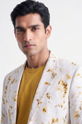 A man wearing a white blazer with a brown and gold floral pattern over a mustard yellow t-shirt, photographed against a clean white background.