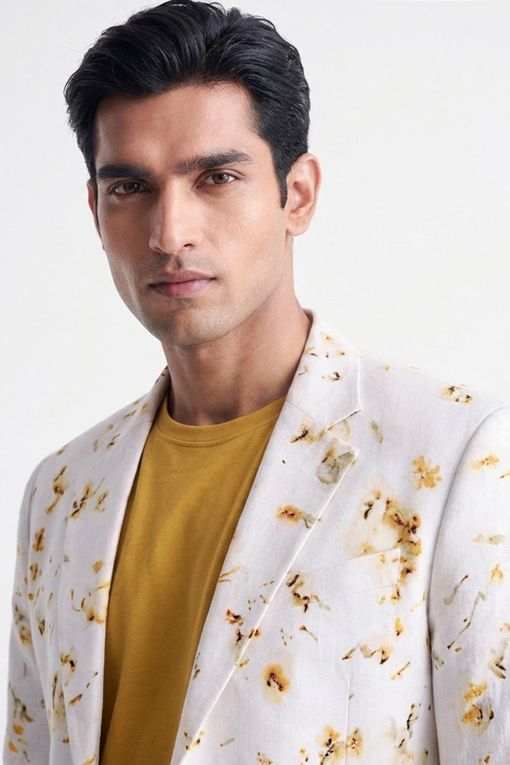 A man wearing a white blazer with a brown and gold floral pattern over a mustard yellow t-shirt, photographed against a clean white background.
