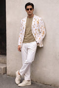 A man wearing a white blazer with a yellow and orange floral pattern, a beige t-shirt, white trousers, and white sneakers, leaning against a textured beige wall.