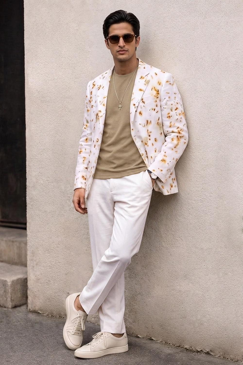 A man wearing a white blazer with a yellow and orange floral pattern, a beige t-shirt, white trousers, and white sneakers, leaning against a textured beige wall.