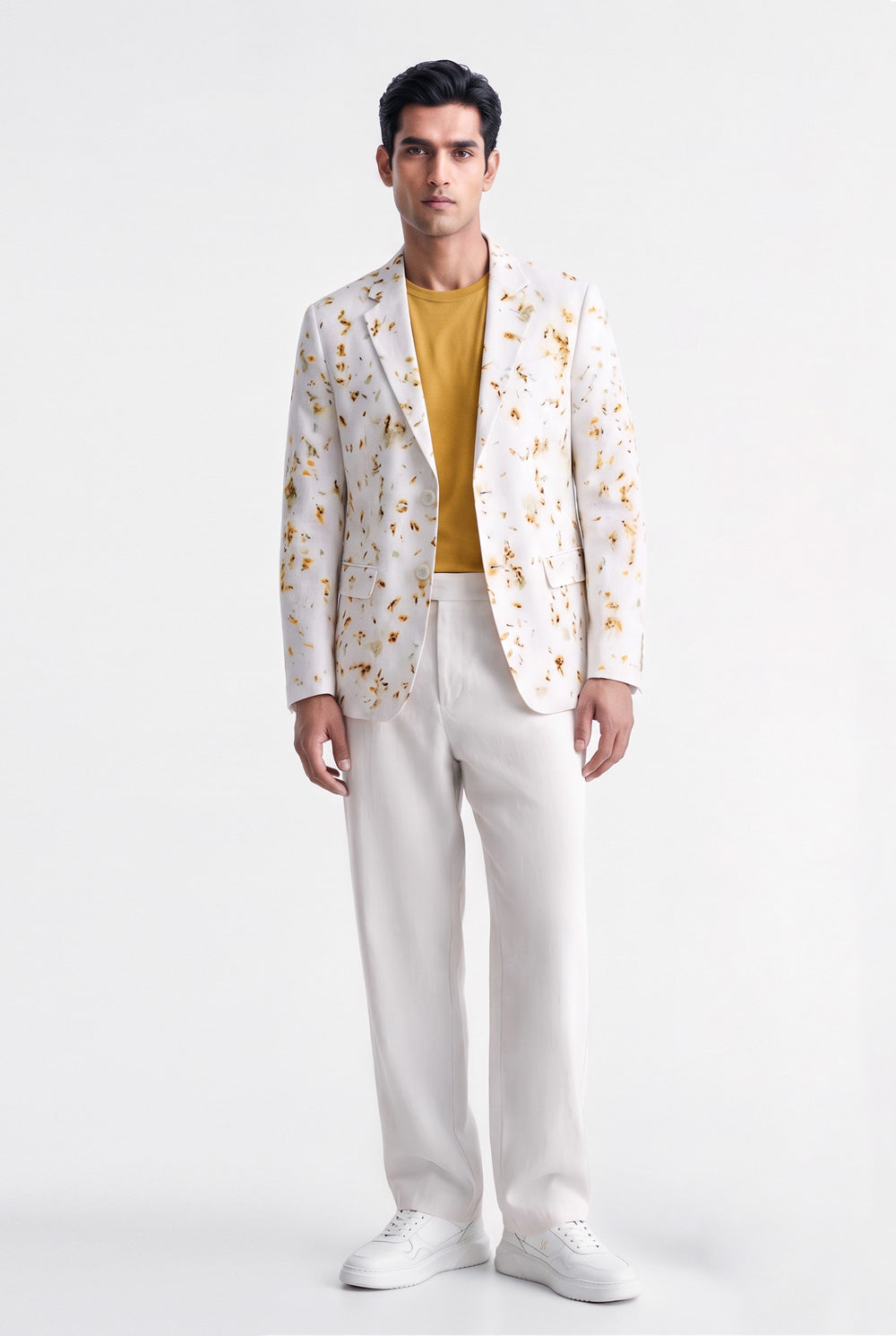 A man modeling a white blazer with a yellow and orange floral pattern, worn over a mustard yellow t-shirt and paired with white trousers and white sneakers, against a plain white studio background.