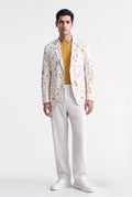 A man modeling a white blazer with a yellow and orange floral pattern, worn over a mustard yellow t-shirt and paired with white trousers and white sneakers, against a plain white studio background.