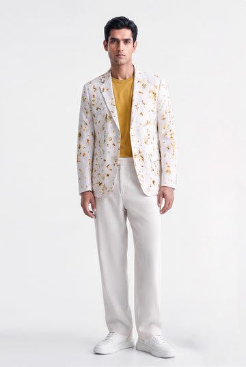 A man modeling a white blazer with a yellow and orange floral pattern, worn over a mustard yellow t-shirt and paired with white trousers and white sneakers, against a plain white studio background.