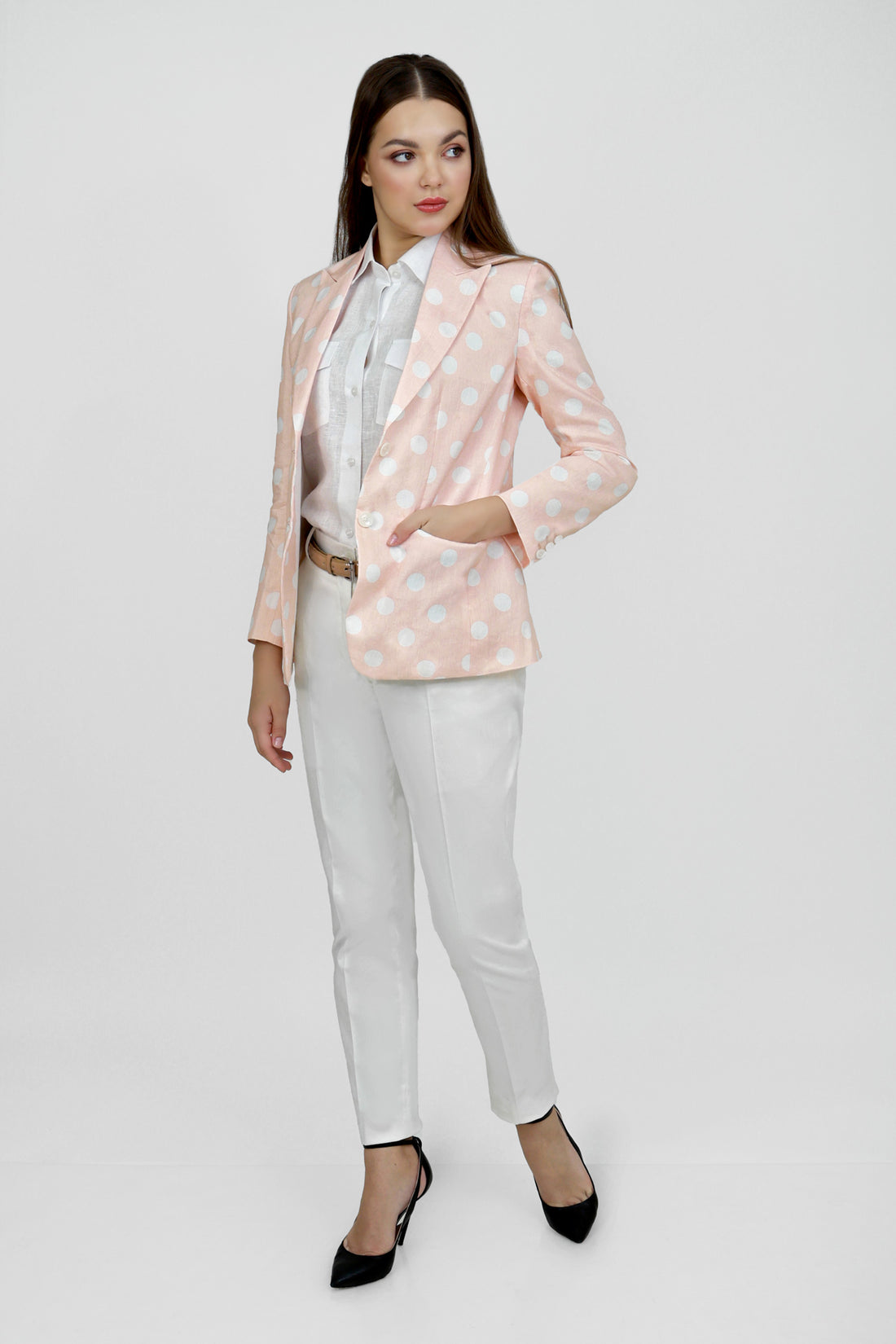 A woman model wearing a peach hemp blazer with a peak lapel, two buttons, and flap pockets, paired with white trousers.