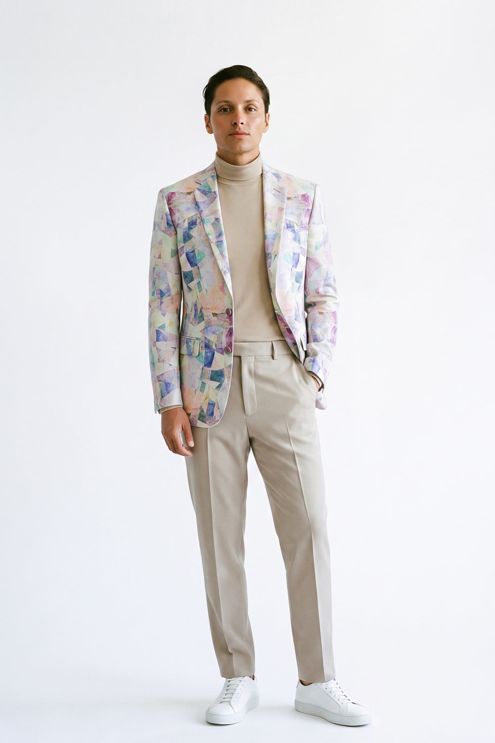 A man modeling a light beige turtleneck sweater, beige trousers, and a patterned hemp blazer with white sneakers, standing against a plain white studio background.