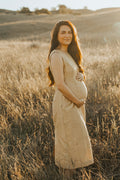 A pregnant woman stands in a sunlit field of dry grass, wearing a sleeveless, knee-length golden silk dress. The warm golden-hour light illuminates the landscape and her silhouette, with rolling hills visible in the background.