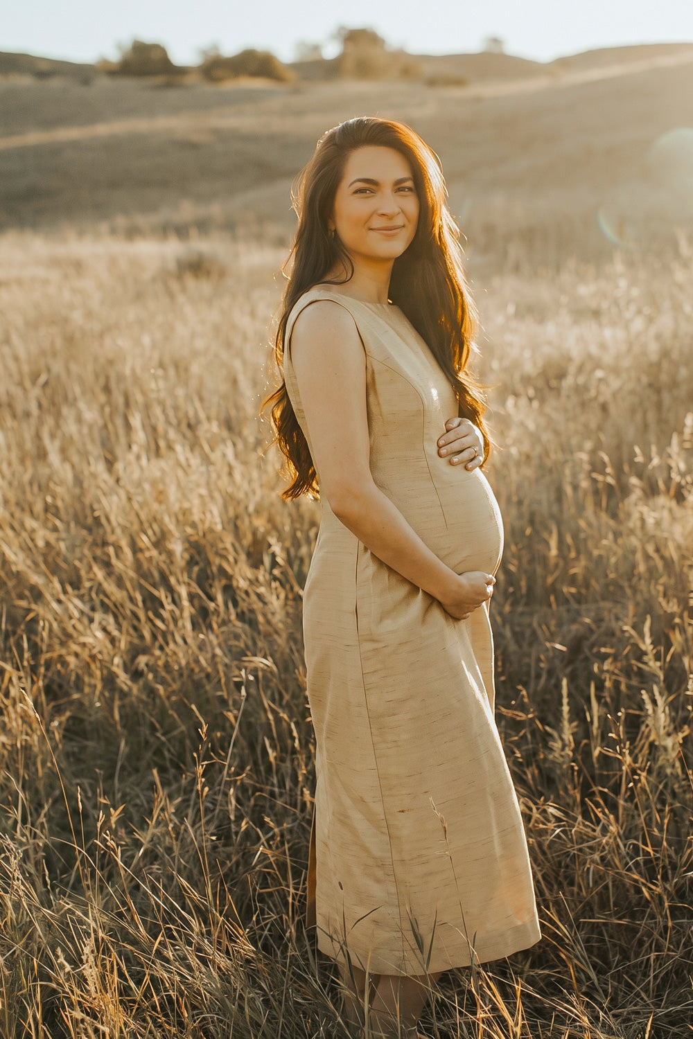 A pregnant woman stands in a sunlit field of dry grass, wearing a sleeveless, knee-length golden silk dress. The warm golden-hour light illuminates the landscape and her silhouette, with rolling hills visible in the background.