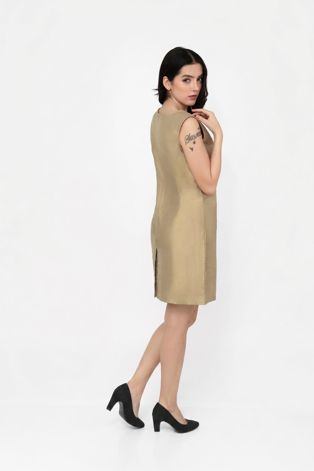 A woman modeling a sleeveless, knee-length beige silk dress with side slits, paired with black heels, photographed against a seamless white studio background.