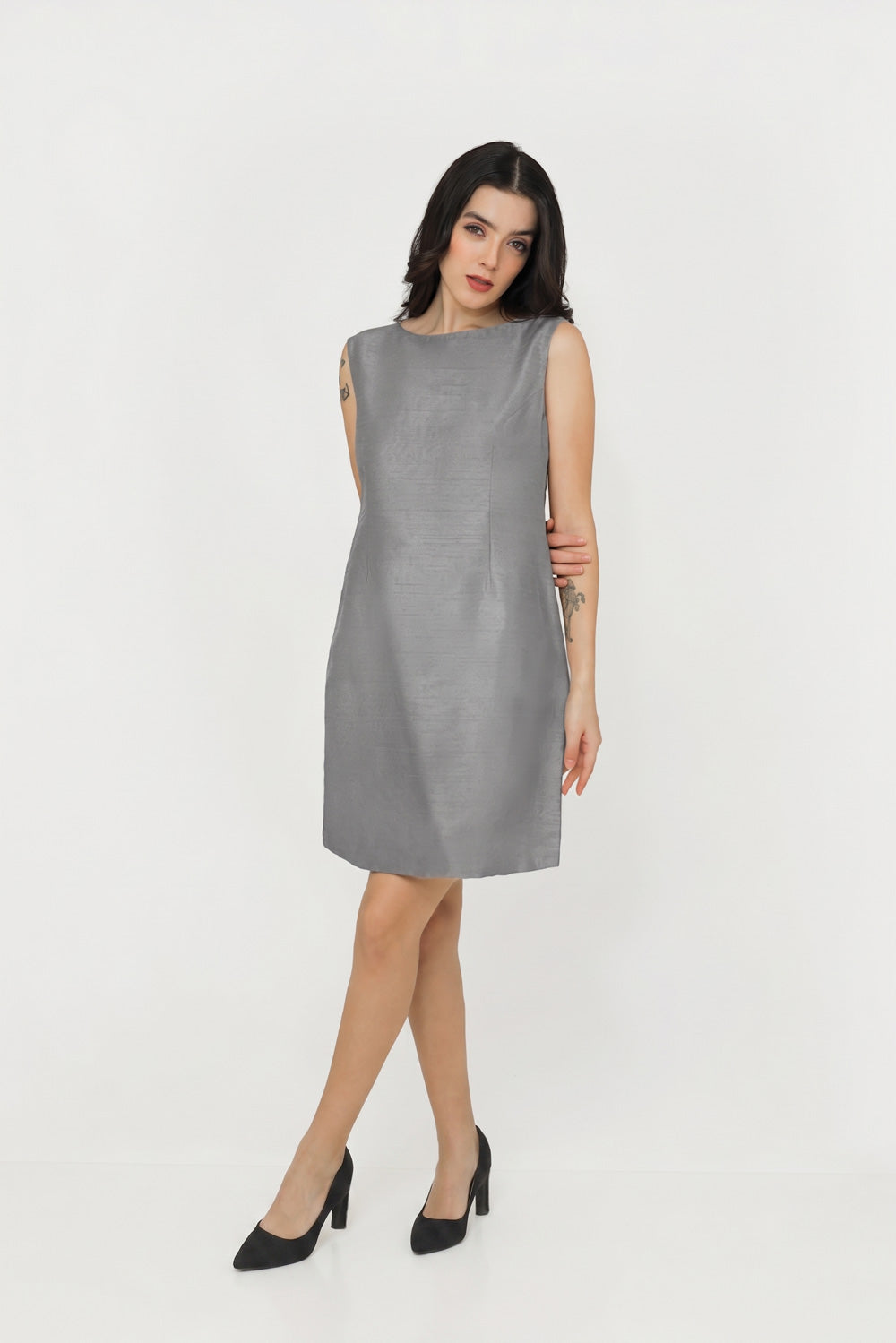 A woman modeling a sleeveless, knee-length gray silk dress with black pointed-toe heels against a plain white studio background.