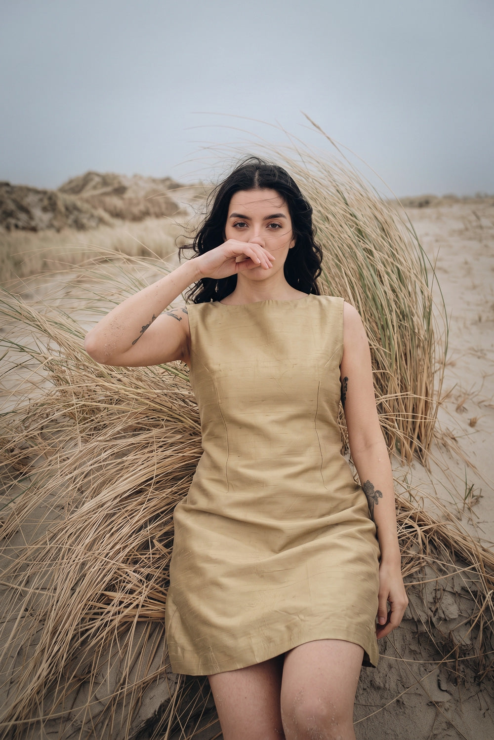 A woman wearing a sleeveless beige silk dress stands on a sandy dune surrounded by tall dry grass under an overcast sky.