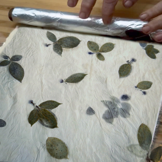 A close-up of hands rolling out silver aluminum foil over a sheet of handmade, crinkled off-white fabric printed with pressed green leaves.