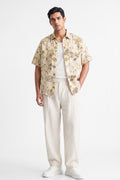 A man modeling a short-sleeve button-up shirt with a beige and brown floral pattern, worn over a white t-shirt and cream-colored linen trousers, against a plain white studio background.