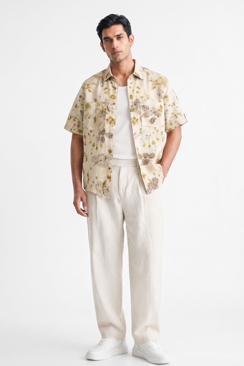 A man modeling a short-sleeve button-up shirt with a beige and brown floral pattern, worn over a white t-shirt and cream-colored linen trousers, against a plain white studio background.