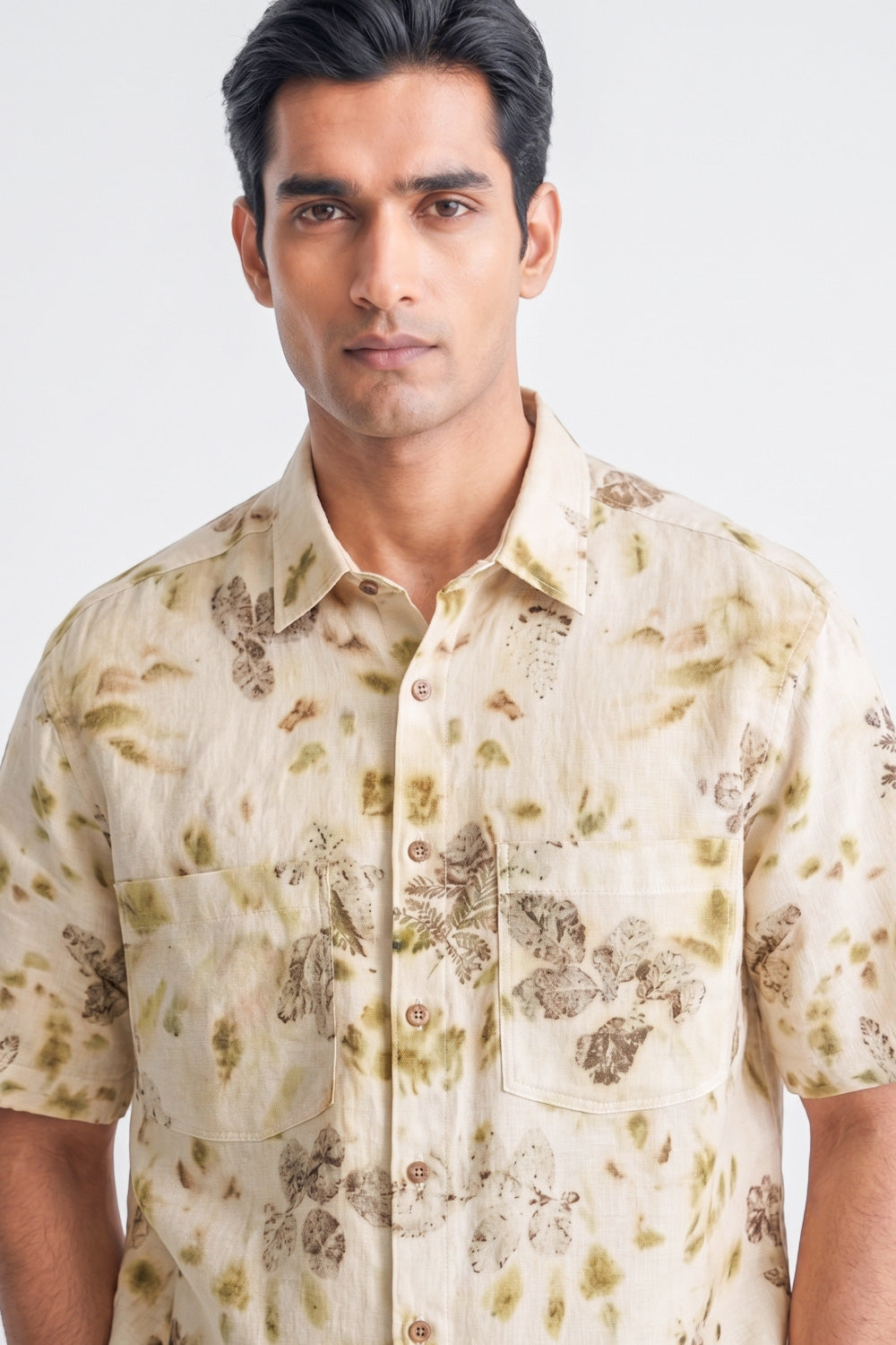 A man wearing a short-sleeve button-up shirt with a beige and olive green floral pattern, photographed against a light gray studio background.