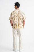 A man viewed from the back wearing a short-sleeve button-up shirt with a beige and brown floral pattern, paired with off-white trousers and white sneakers, against a plain white studio background.
