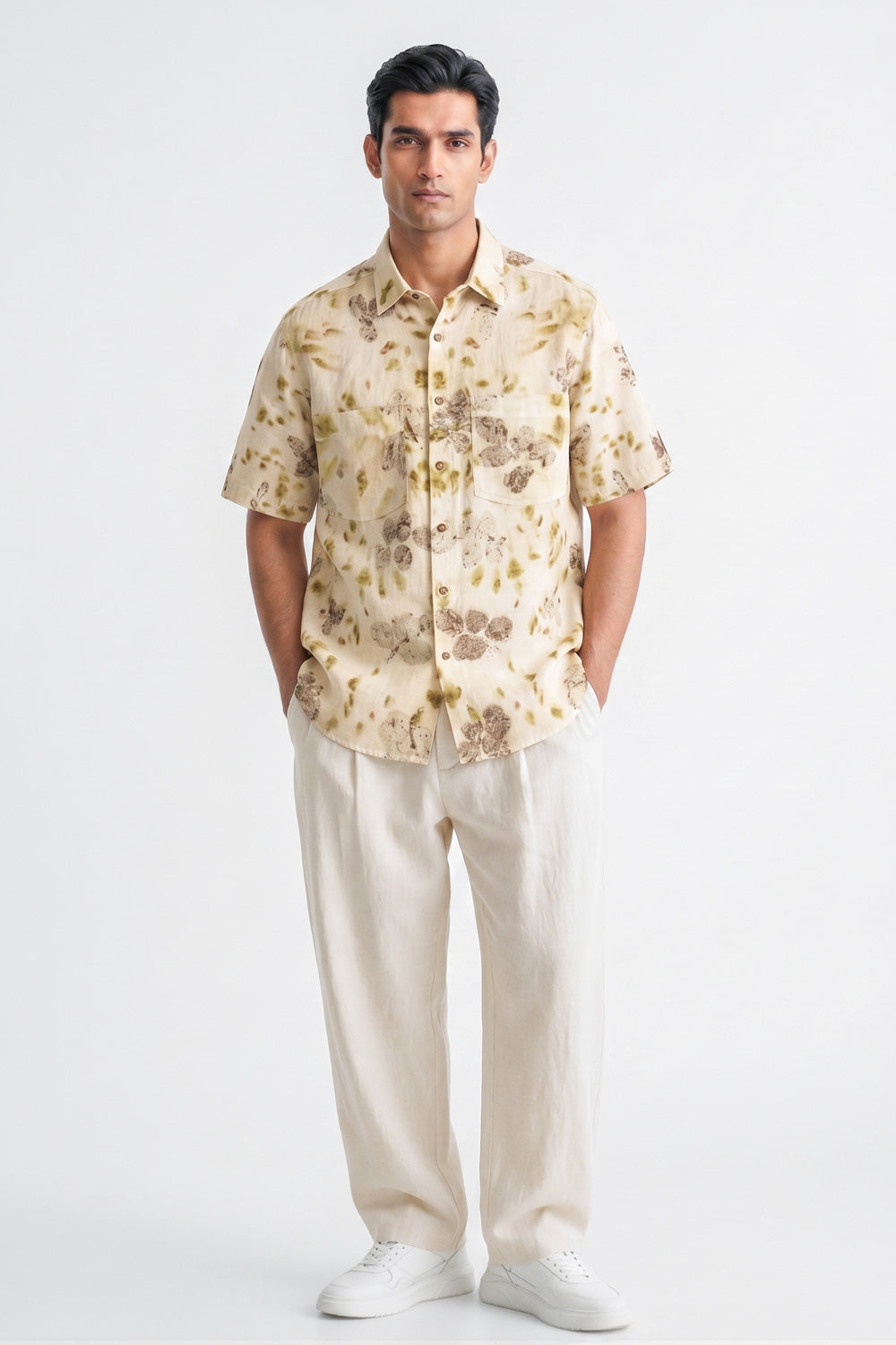 A man modeling a short-sleeve button-up shirt with a beige and brown floral pattern, paired with cream trousers and white sneakers, against a plain white studio background.