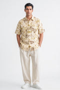A man modeling a short-sleeve button-up shirt with a beige and brown floral pattern, paired with cream trousers and white sneakers, against a plain white studio background.