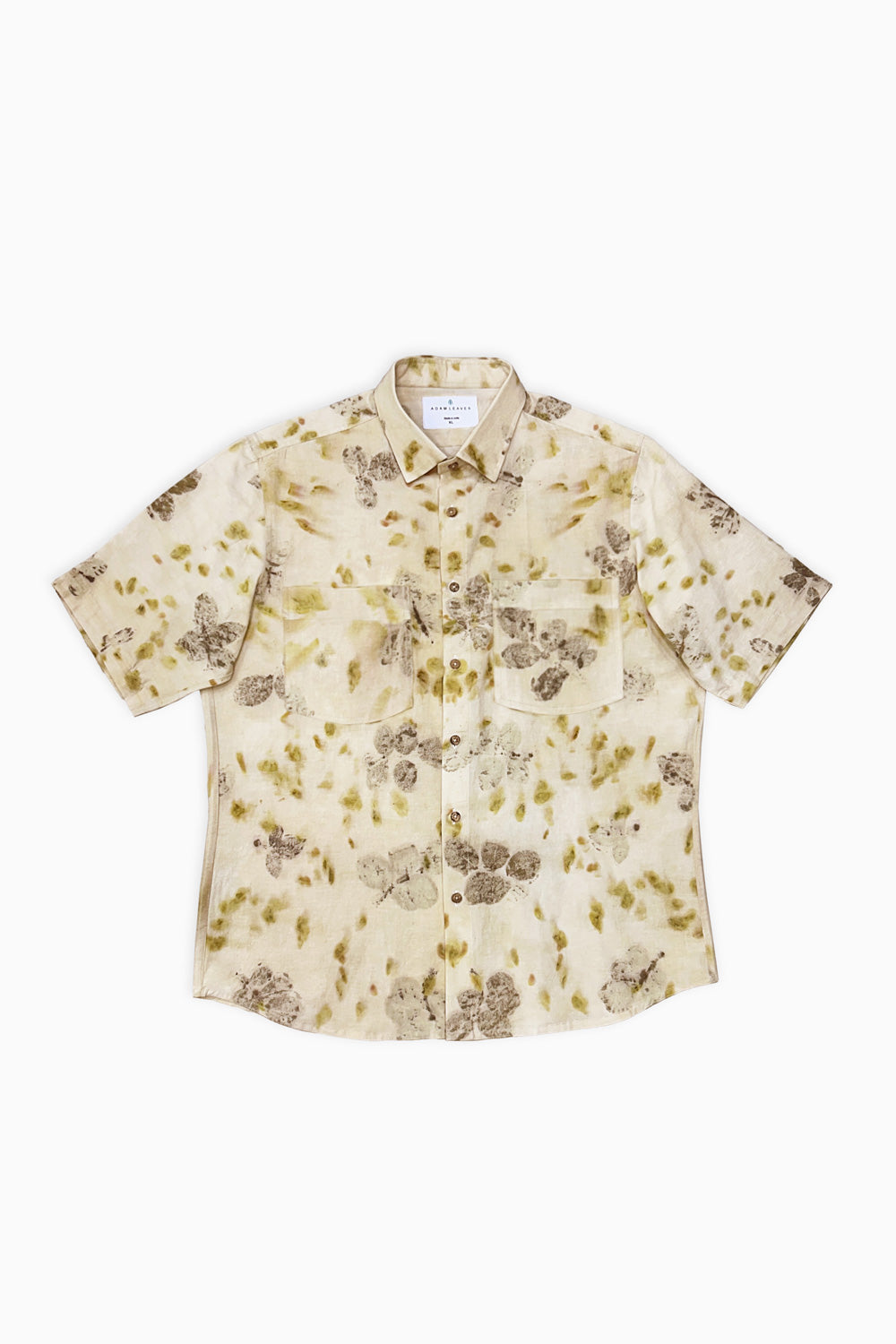 A man modeling a short-sleeve button-up shirt with a beige and brown floral pattern, paired with cream trousers and white sneakers, against a plain white studio background.