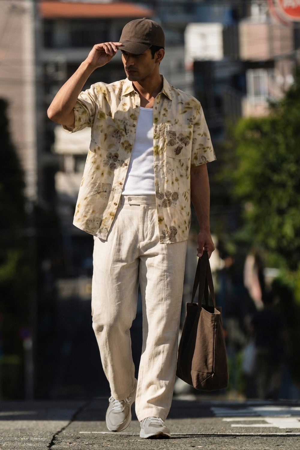 A man walking outdoors on a city street wearing a beige short-sleeve button-up shirt with a muted floral print, cream trousers, a white tank top, a brown cap, and carrying a brown tote bag.