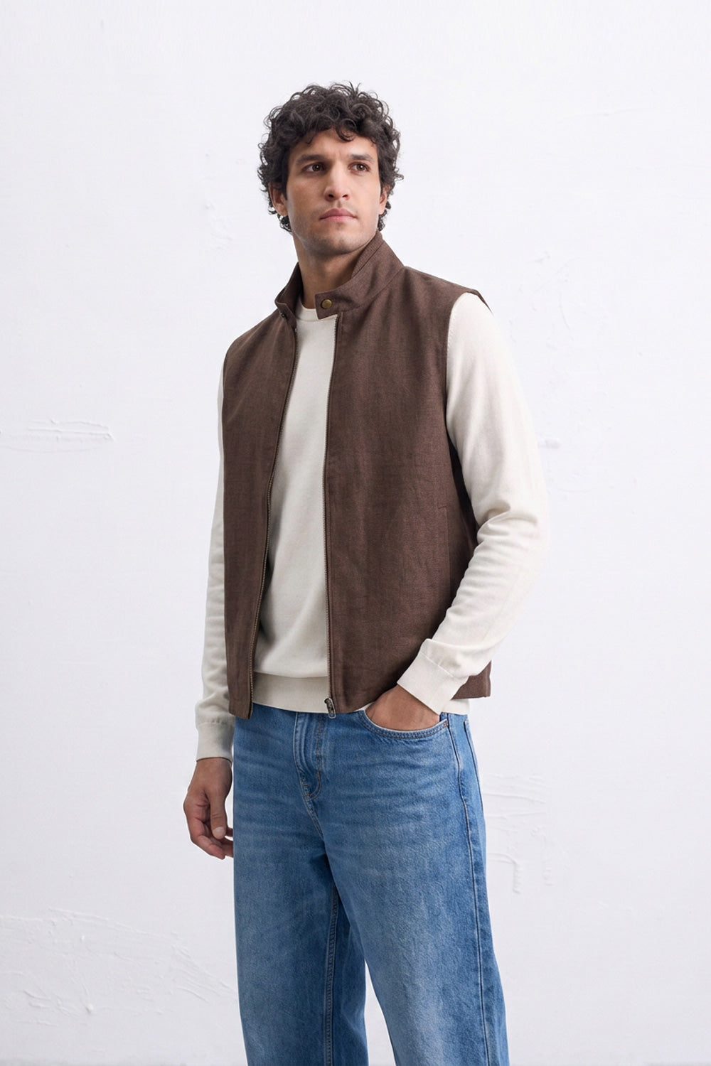 A man wearing a brown sleeveless zip-front linen vest over a cream long-sleeve shirt and blue jeans, standing against a white wall.