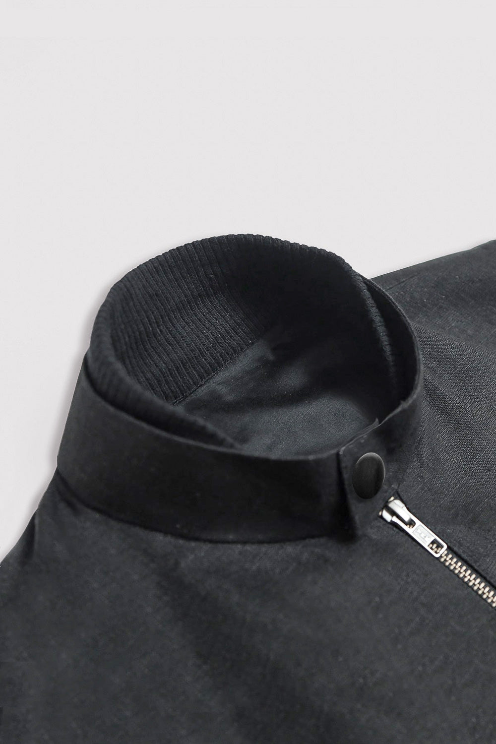 Close-up of a black linen jacket with a ribbed knit stand collar, snap button, and a partial silver zipper on a light gray background.