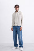 A man modeling a light beige zip-up linen jacket over a matching beige sweater, paired with blue jeans and white sneakers, standing against a white studio background.