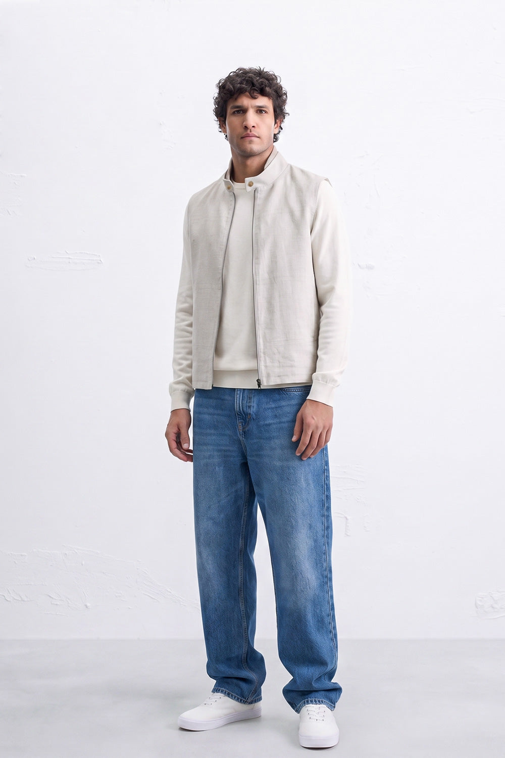 A man modeling a light beige zip-up linen jacket over a matching beige sweater, paired with blue jeans and white sneakers, standing against a white studio background.