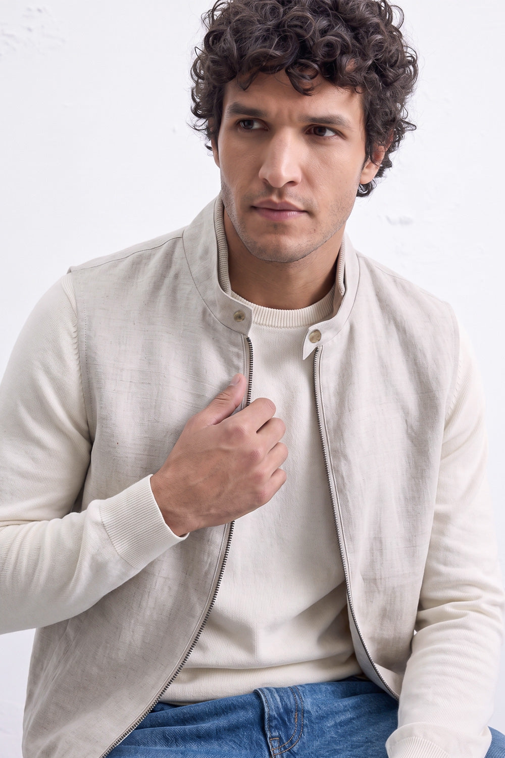A man wearing a light beige linen zip-front jacket over a cream crewneck sweater and blue jeans, posed against a white textured wall.