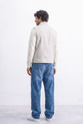 A man viewed from behind wearing a cream-colored knit sweater and linen vest, blue jeans, and white sneakers, standing against a white textured wall.