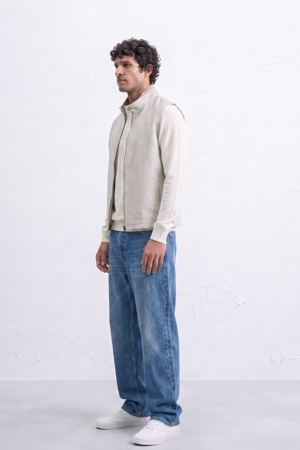 A man wearing a light beige zip-up linen jacket over a white shirt, blue jeans, and white sneakers, standing in profile against a white textured wall.