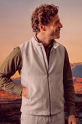 A man wearing a light beige zip-up linen vest over an olive green long-sleeve shirt, standing outdoors against a warm sunset sky with mountains in the background.