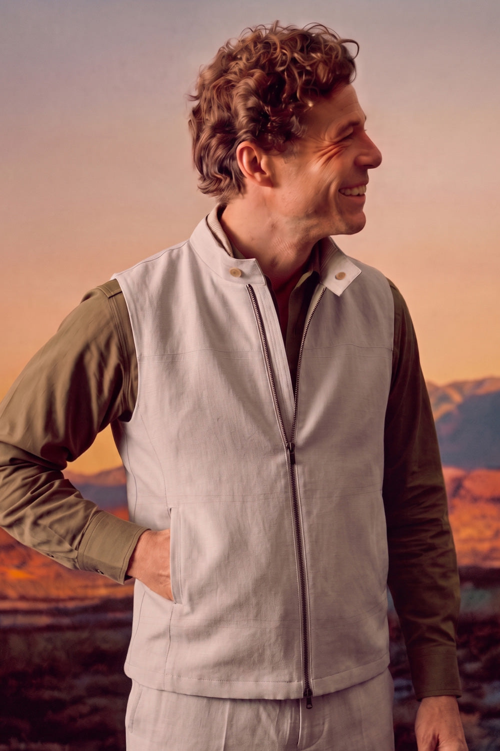 A man wearing a light beige zip-up linen vest over an olive green long-sleeve shirt, standing outdoors against a warm sunset sky with mountains in the background.