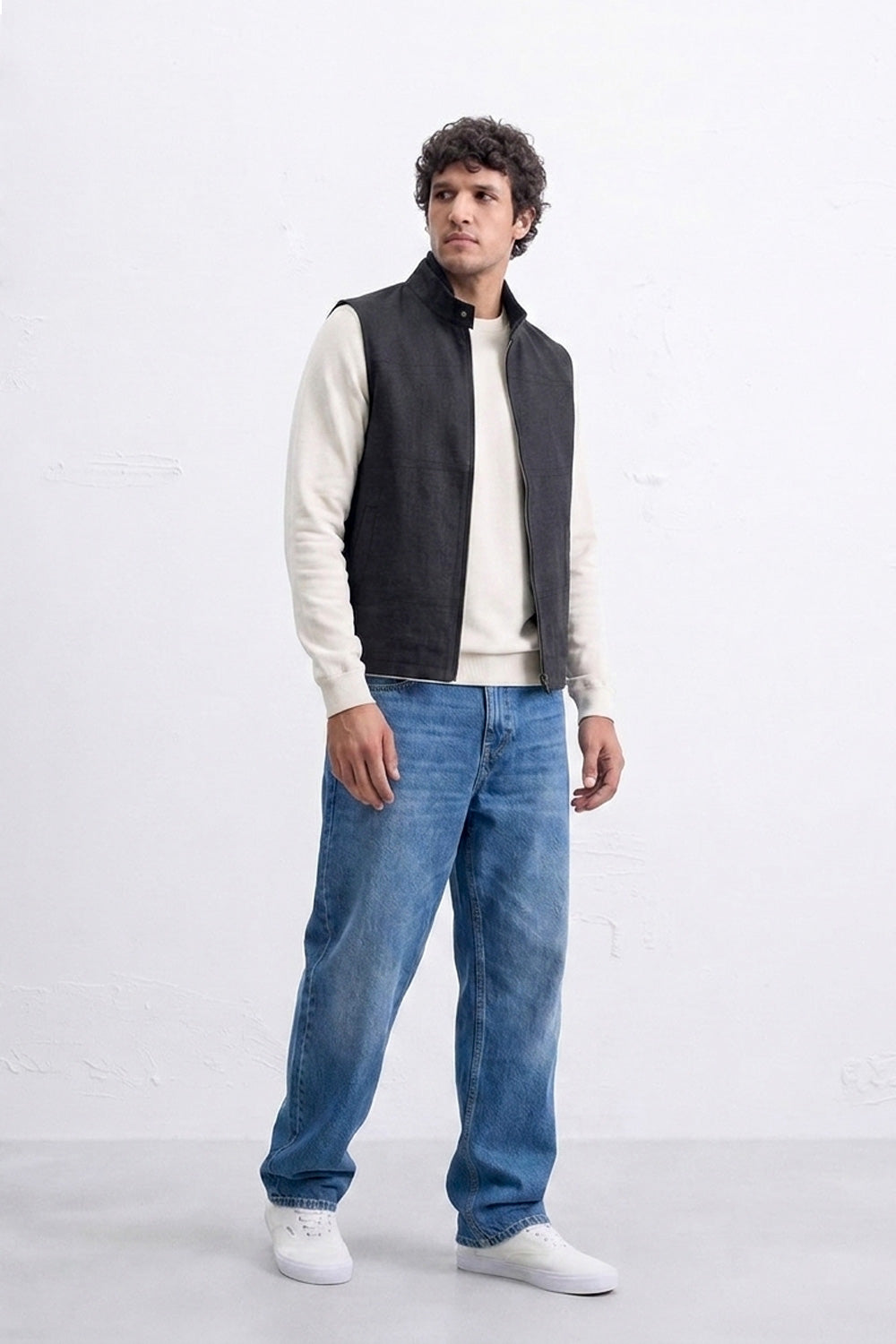 A man modeling a black sleeveless zippered linen vest over a cream long-sleeve shirt, paired with blue jeans and white sneakers, standing against a white studio wall.
