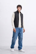 A man modeling a black sleeveless zippered linen vest over a cream long-sleeve shirt, paired with blue jeans and white sneakers, standing against a white studio wall.