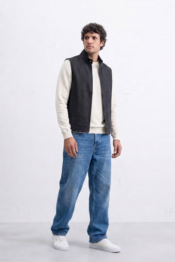 A man modeling a black sleeveless zippered linen vest over a cream long-sleeve shirt, paired with blue jeans and white sneakers, standing against a white studio wall.