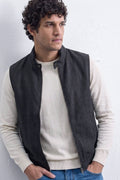A man wearing a dark charcoal gray sleeveless zip-front linen vest over a cream knit sweater and blue jeans, standing against a white wall.