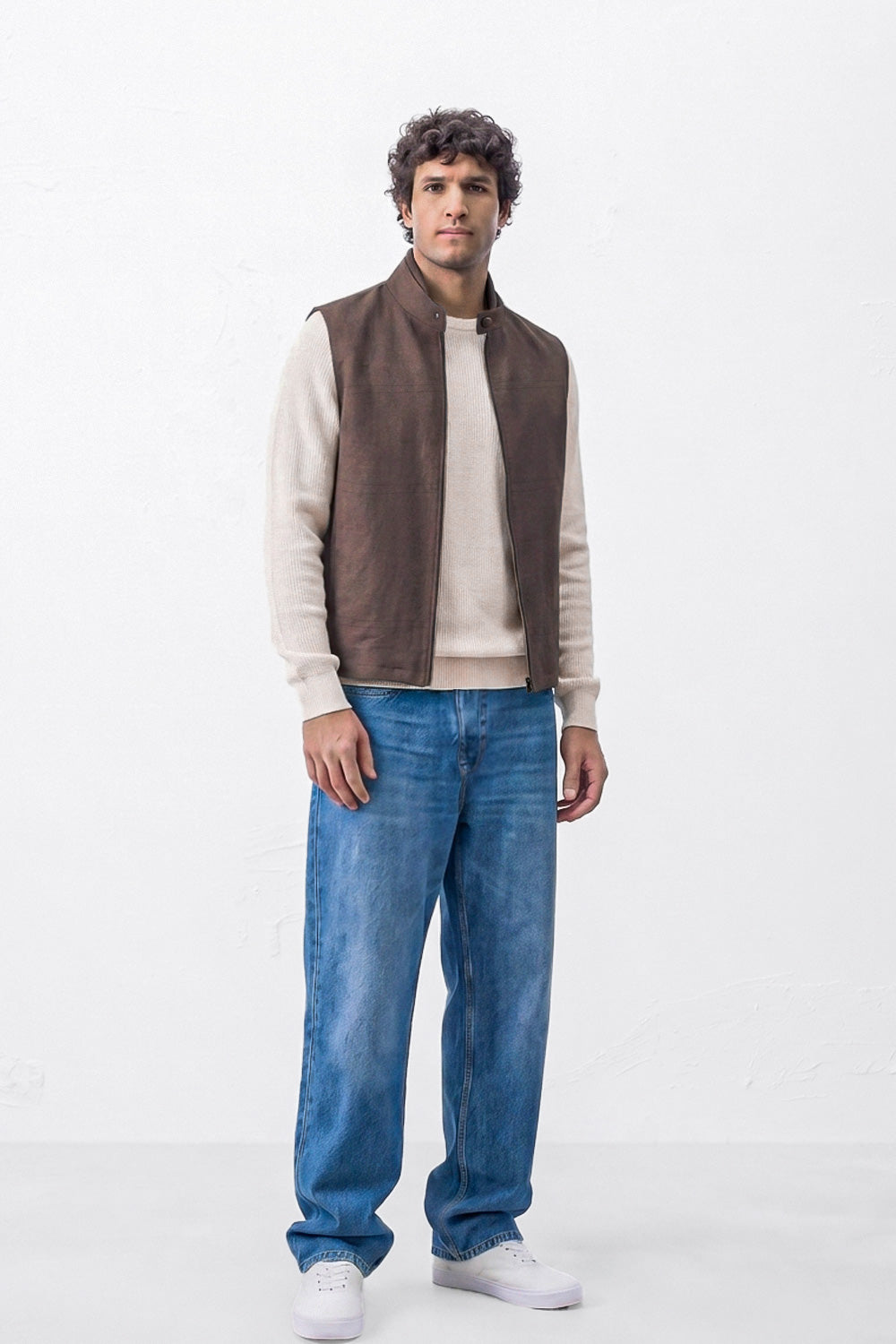 A man modeling a brown linen sleeveless vest over a beige long-sleeve shirt, paired with blue jeans and white sneakers, standing against a plain white studio background.