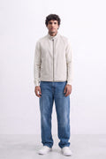 A man modeling a light beige zip-up linen jacket with a stand collar, paired with blue jeans and white sneakers, standing against a white studio background.