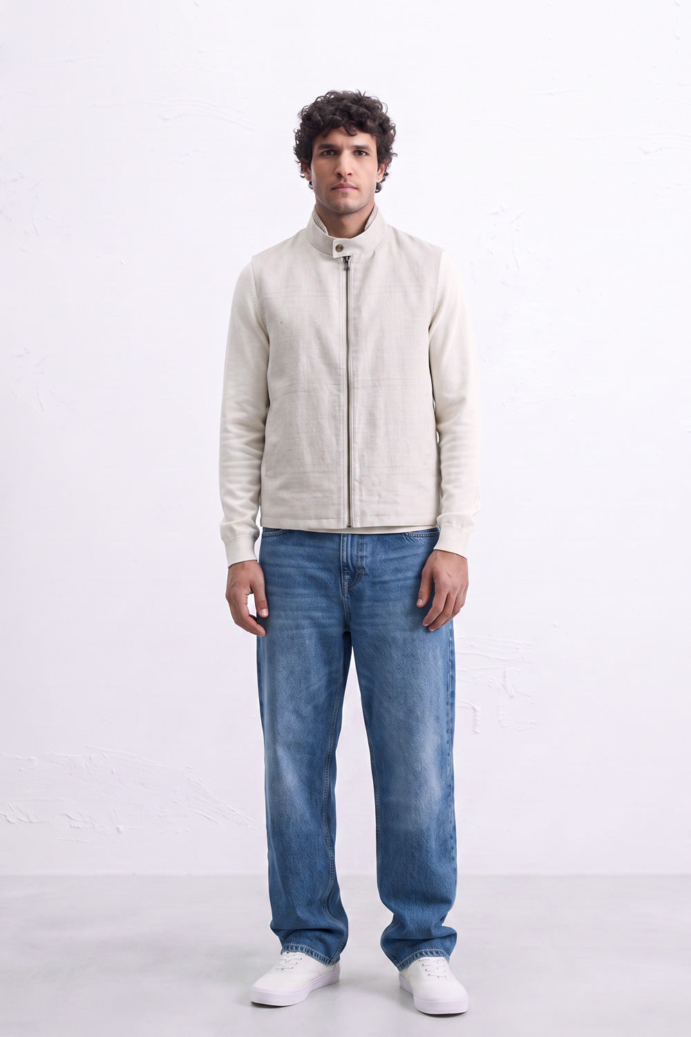 A man modeling a light beige zip-up linen jacket with a stand collar, paired with blue jeans and white sneakers, standing against a white studio background.