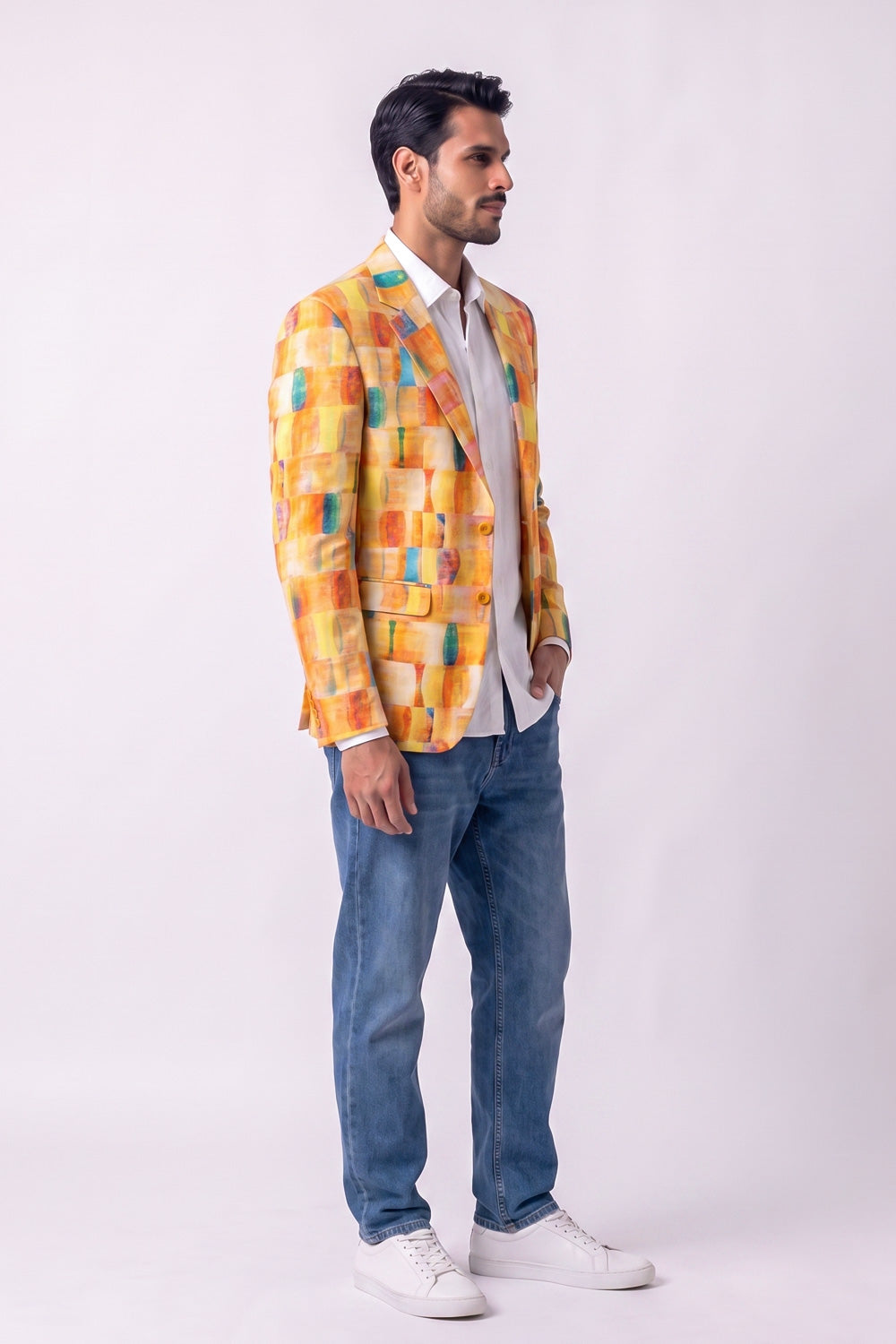 A man wearing a vibrant yellow, orange, and blue geometric patterned hemp blazer over a white shirt, paired with blue jeans and white sneakers, standing against a plain white studio background.