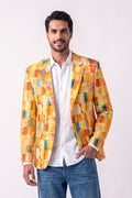A man wearing a vibrant yellow hemp blazer with a colorful abstract geometric pattern, paired with a white button-down shirt and blue jeans, standing against a plain white studio background.