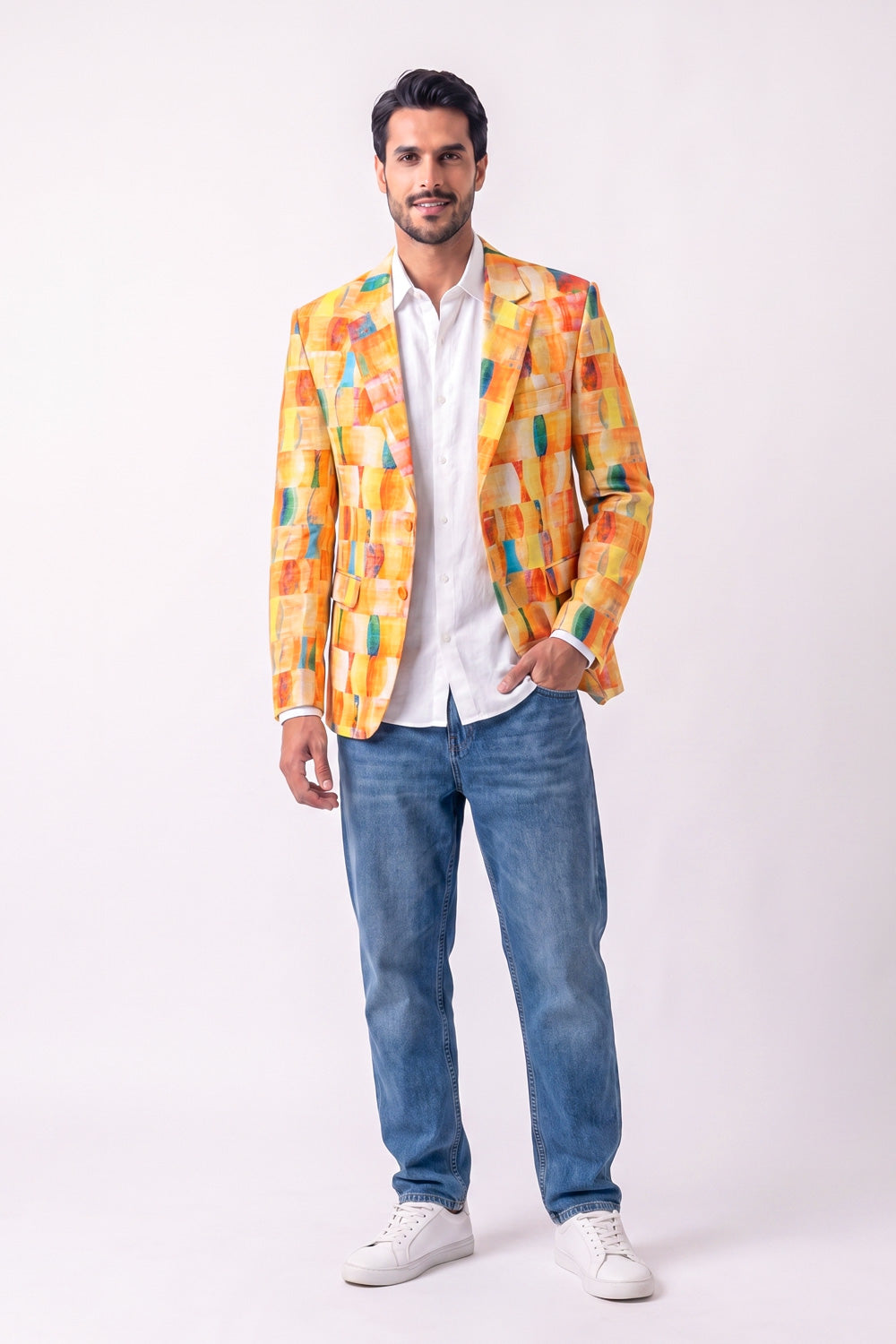 A man modeling a vibrant yellow, orange, and green abstract-patterned hemp blazer over a white button-down shirt, paired with blue jeans and white sneakers, against a plain light background.