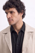 A man with curly dark hair wearing a light beige linen over-coat over a black button-down shirt, photographed against a plain white background.