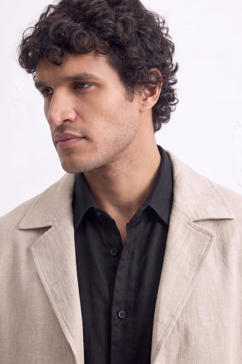 A man with curly dark hair wearing a light beige linen over-coat over a black button-down shirt, photographed against a plain white background.